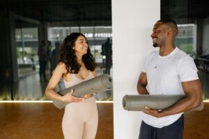 A smiling man and woman holding yoga mats, preparing for a fitness class in a modern gym.